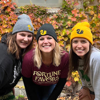 Three people wearing Fortune Favors beanies in gray, black and gold